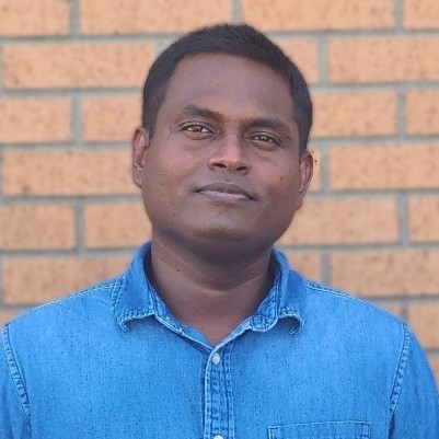 Man in collared denim shirt half smiling, brick wall background