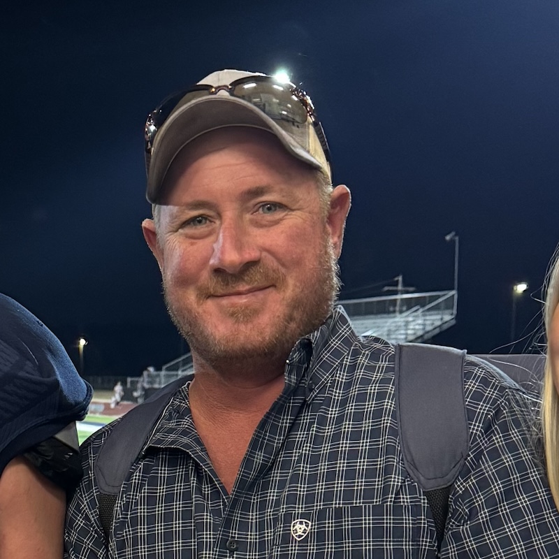 Man with tan baseball cap, sunglasses resting on cap, with navy plaid button up shirt, and light facial hair, smiling